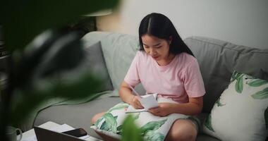 Footage of Young Asian woman checks her finances using a calculator on smartphone and makes notes in a notebook while sitting on the sofa at home,Home finance video