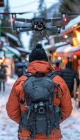 A man wearing outerwear with a backpack, with a drone flying above him photo