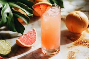 A highball glass filled with a refreshing grapefruit iced beverage, garnished with a grapefruit wedge. Surrounding are fresh grapefruits, lime, and monstera leaves, all set on a marble surface. photo