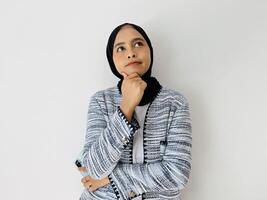 Portrait of Thoughtful Asian Muslim woman standing with her hand on chin thinking, feeling doubtful and confused, with different options, wondering which decision to make while holding phone photo