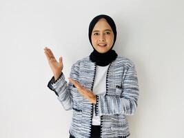Smiling young Asian woman wearing head wearing black hijab and tweed blazer showing and pointing something standing over white background. Copy space for your text photo