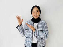 Smiling young Asian woman wearing head wearing black hijab and tweed blazer showing and pointing something standing over white background. Copy space for your text photo
