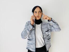 An Asian woman wearing hijab thinks deeply about something while answering phone, uses modern mobile phone, keeps index fingering on head isolated by white background photo