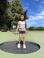Little Asian child enjoys jumping on trampoline. Happy little girl bouncing on a trampoline on the playground in a park in the afternoon. Outdoor activity photo