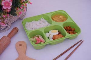 A green tray with five compartments containing white rice, curry, sliced cucumbers and tomatoes, a boiled egg, quail eggs, fried chicken, and pieces of meat photo