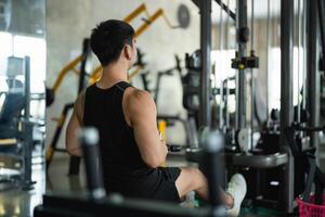 Back view. Asian man in a black tank top works out on a seated row machine in a modern gym. His serious expression and muscular build emphasize his dedication to fitness and strength training. photo