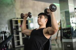 Asian man in a black tank top performs a seated dumbbell shoulder press in a modern gym. His muscular build and concentrated expression highlight his dedication to strength training. photo