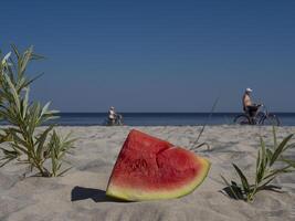 A piece of red watermelon on sand near sea and blue sky in background with two people riding bicycle. The scene showcases refreshing slice of watermelon on beach while cyclists pass by. photo