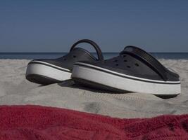 Black and white beach slippers stand on sand near the sea, with red towel in front. Slippers set by the water's edge, waiting for day at beach. Peaceful scene with beach slippers, sand, and ocean. photo