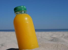 Orange juice in a plastic bottle with green lid stands in the sand on the seashore. The scene features orange juice as a refreshing companion for relaxing beach day Concept Healthy Nutrition Vitamins. photo