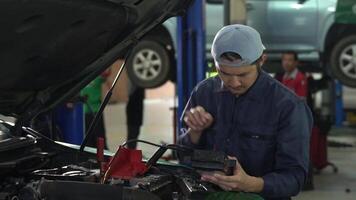 Asian auto mechanic checking battery voltage maintenance With digital battery tester showing thumb up and looking at camera in garage car service video