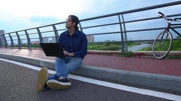 Asian man sitting on the side of the road with an open laptop in front of him video