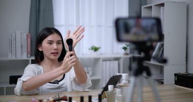 Handheld shot, Beautiful asian woman blogger is showing how to makeup and use cosmetics. In front of the smartphone to record vlog video live streaming at home. Influencer and cosmetics concepts.