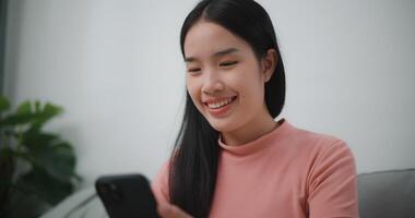 Footage of Happy young asian woman relax on comfortable couch at home shopping online on smartphone, smiling girl use cell phone chatting, browse wireless internet on gadget video