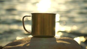 Close-up of a travel stainless mug standing on a rock. Small waves on golden warm water surface with bokeh lights from sun. A concept of the world of beauty, nature and outdoor travel. Selective Focus video