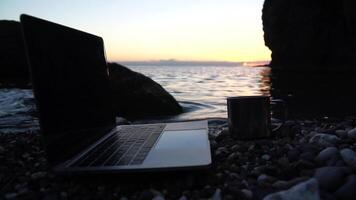 Silhouette of laptop and cup on the beach over sea sunset. Sparkling sea water on background. Freelancer, blogger working on notebook on the beach. video