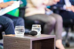two glasses of water are prepare to speakers who sit on the stage with microphone beside it. photo