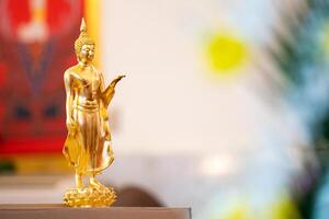 Standing Buddha statue is placed on top of a rectangular object in front of a red framed picture and tree house. photo