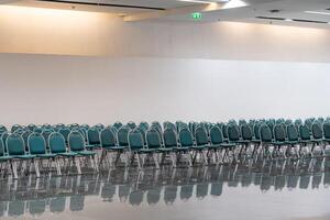 Many empty green leather chairs in a wide hall room, lights on. A floor reflect the objects. Give a feeling of emptiness. photo