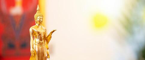 Standing Buddha statue is placed on top of a rectangular object in front of a red framed picture and tree house. photo