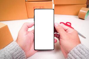 Goods delivery concept , mobile phone with blank white screen in a human hand and cardboard boxes on desk for product packing , copy space for text photo