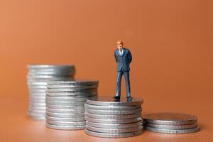 Miniature people A businessman in a suit stands with several stacks of silver coins photo