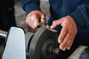 hands of grinder male sharpener sharpening a knife blade on a grinding machine with abrasive wheel in the workshop close-up photo