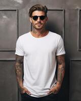 A handsome man in a white t-shirt stands confidently in front of a wall photo