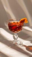 A glass of red cocktail with an orange twist sits on a white surface bathed in sunlight photo