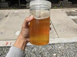 Hand holding a jar of industry oil sample. A close up of a hand holding a glass jar filled with a yellow liquid and some foam, with a man standing in the background. photo