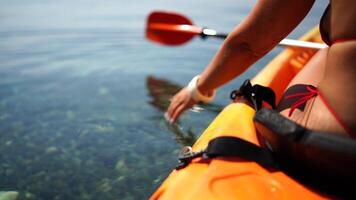 A woman is sitting in a kayak on a lake. The kayak is orange and the paddle is red. The woman is wearing a bikini and she is enjoying her time on the water. video