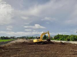 West Lombok Regency, Indonesia - February 4, 2024. Outdoors, a Backhoe is at work leveling the ground for a housing site, with a blue sky in the background. photo