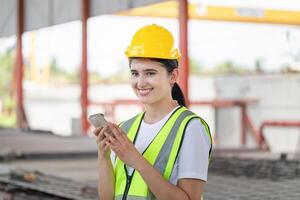 alegre hembra ingeniero en el difícil sombrero usos móvil teléfono, industrial trabajador utilizando móvil teléfono inteligente en construcción sitio foto