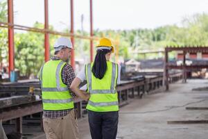 espalda ver de ingeniero y capataz trabajador equipo con planos comprobación proyecto a el prefabricado hormigón fábrica sitio, ingeniero y constructores en sombreros duros que se discute en construcción sitio foto