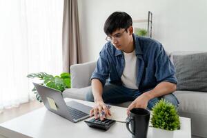 A man is sitting on a couch and working on a laptop. He is using a calculator and a pen photo