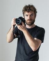 Man with tousled brown hair and a beard is holding a camera looking directly at the viewer photography photo