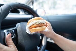 Asian lady holding hamburger to eat in car, dangerous and risk an accident. photo