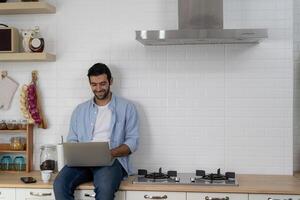 Freelancer working from home sitting on a kitchen worktop and using laptop. Bearded man working with a laptop and reading news. photo