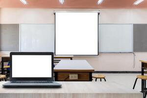 Laptop on table foreground. Projector screen canvas in modern conference room with big windows. Side view. Empty classroom or presentation room interior with desks photo