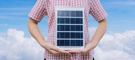 Successful woman holding solar panels and wind turbine with folded arms, isolated on blue sky background. Clean system technology and safety photo