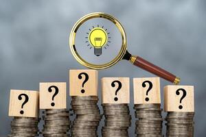 A magnifying glass with a light bulb icon and a question mark on a wooden block resting on a stack of coins has an idea that can solve financial problems. photo