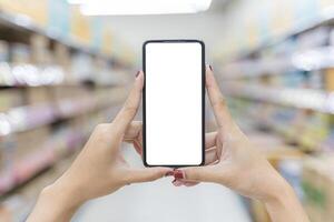 Close up of two female hands holding a mobile phone on a vertical blank white screen. Blurred supermarket background. Mockup. photo