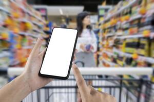 A person's hand holds a mobile phone, with a shopping basket on the screen. In the background, a pretty woman selects digital products, in a blur. photo
