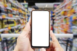 A person's hand holds a mobile phone, with a shopping basket on the screen. In the background, a pretty woman selects digital products, in a blur. Mock up. photo