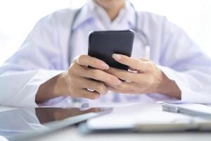 Female doctor hands holding and using mobile smart phone with laptop computer and stethoscope on the table photo
