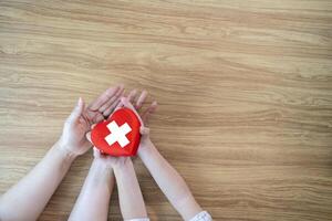 Red Heart with cross in hands of the child and mother on a white wooden photo