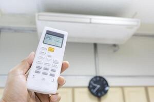 The hand that is holding the remote to control the air conditioner requires a cooler temperature. The background is an air conditioner that is running at full capacity. photo