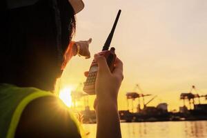 Caucasian female foreman is talking with colleague by using walkie talkie and standing in front of industrial containers. Engineering and Technology concept. photo