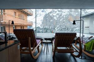 Interior of modern space with mountain view through large window. Wooden rocking chairs, black side table, floor lamp, snowy peaks. Serene atmosphere. Location Unknown photo
