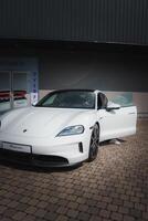 White Porsche Taycan parked in front of a modern building. Driver side door open with person inside. Clean, sleek luxury car design. photo